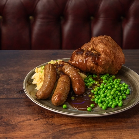 A plated serving of Cumberland sausage and Colcannon Mash with a ladle pan of gravy, Yorkshire Pudding and peas, on a table.