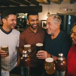 A group of friends stood round the bar area drinking pints in a Greene King venue.