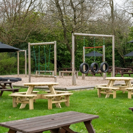The exterior beer garden seating area and children's play area at The Hussey Arms in Brownhills, with wooden picnic tables, shade umbrellas, and obstacle course.