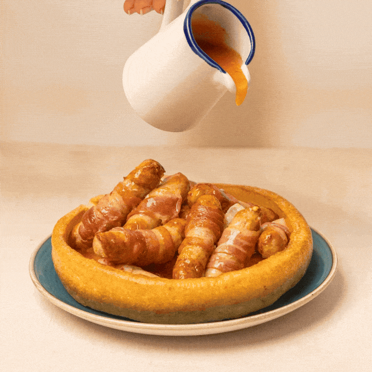 A GIF of sauce being drizzled from a jug over pigs in blankets which are served in a giant Yorkshire pudding on a plate.