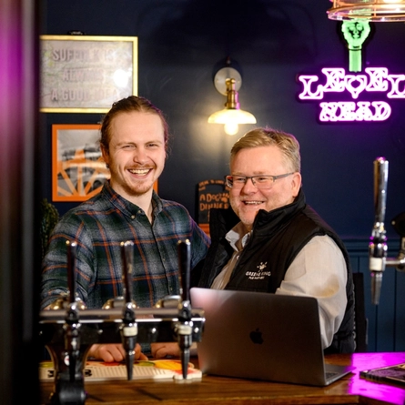 Two Men stood at a bar smiling with a laptop in front of them