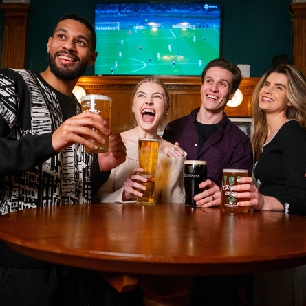 An image of a group of friends stood at an interior table enjoying drinks while watching a football match shown on TV within an Urban Social venue.
