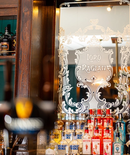 A mirror engraved with the words "Lord Raglan" and filigree patterns, and bottles of whisky and other alcoholic spirits on wooden shelves, behind the bar at the Lord Raglan.