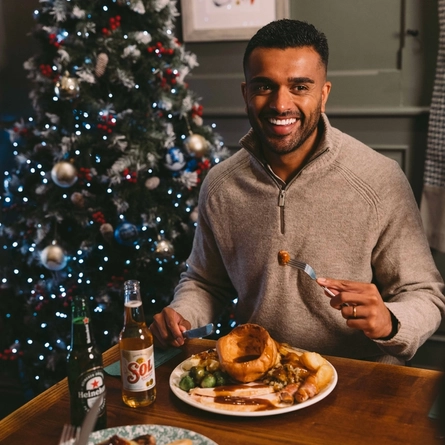 Person seated at a festive dining table holding a forkful of food above a traditional Christmas roast dinner with Yorkshire pudding, roast potatoes, vegetables and gravy. Beer bottles sit on the table, and a decorated Christmas tree glows in the background.