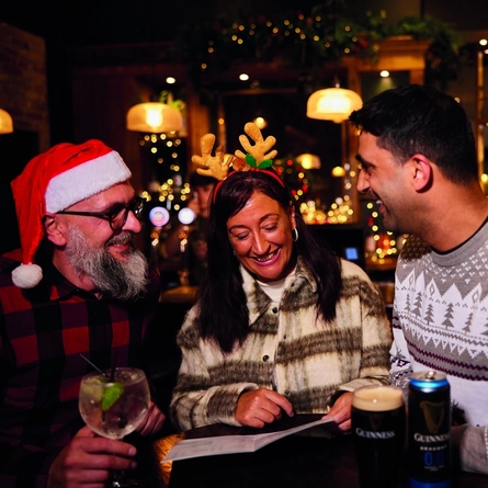 Three people are standing at a tall table with drinks,  in a festive, warmly lit bar. Two of them are wearing Christmas themed headwear.  The background features Christmas decorations, including garlands and lights, as well as hanging glasses above the bar area.