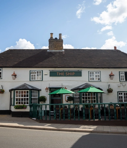 A view from across the road of the exterior facade, signage, and seating area of The Ship in Bedford; with shade umbrellas on some of the tables, and flower baskets and golden lanterns hanging on the white walls of the pub.