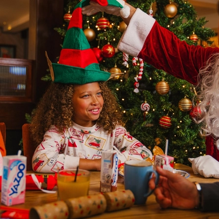 A child wearing Christmas-print pyjamas sits at a festive breakfast table while a person dressed as Santa Claus places a green-and-red elf hat on their head. A decorated Christmas tree with red and gold baubles, candy cane ornaments and warm fairy lights stands behind them. The table is set with breakfast dishes, juice cartons, a glass of orange juice, mugs, Christmas crackers and paper party hats, with other diners seated around the table in a Christmas-themed restaurant setting.