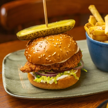 A close up view of a Chicken Burger served on a plate with a small bowl of fries. The plate sits on a wooden restaurant table next to another plate of food.