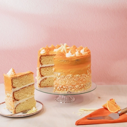 The Simply The Zest cake on a cake stand, on a table next to a slice of the cake served on a plate, some orange segments, and a cake slicer. 