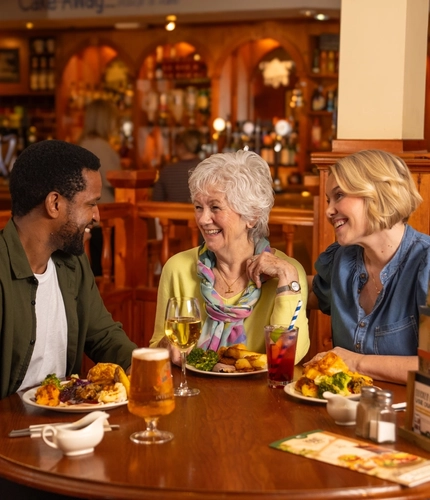 Three people sitting at a wooden restaurant table with plates of roast dinner in front of them. A glass of wine, a glass of beer, and a glass of fizzy drink also sit on the table, along with menus, salt, pepper, and jugs of gravy.