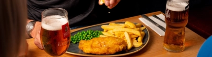 A hand squeezing a lemon wedge over a plated portion of fish and chips, at an indoor table.