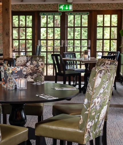 A close up view of a circular wooden table in the interior restaurant seating area of The Hollybush in Lichfield, with upholstered chairs around the table, and wooden beams and floor to ceiling windows visible in the background  
