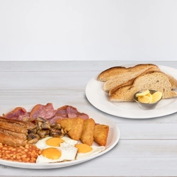 The mega breakfast meal at Hungry Horse venues, featuring sausages, bacon, mushrooms, hash browns, fried eggs and baked beans served on a plate. The plate sits on a table next to a plate of sliced bread and a small pot of butter.