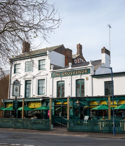 A view from across the road of the exterior facade, signage, and seating area of The Grosvenor in Felixstowe, with shade umbrellas on the tables, and string lights above them.