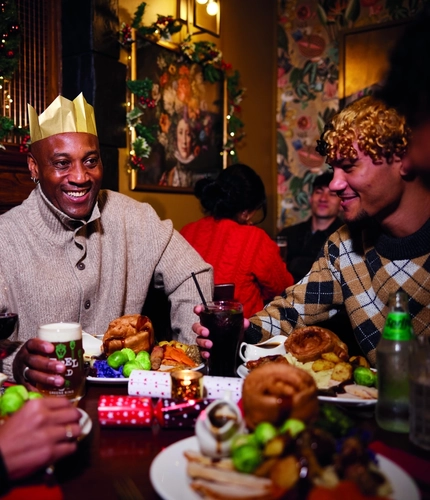 A group of people are gathered around a table enjoying a festive meal. They are wearing paper crowns, and the table is set with plates of food. The background is decorated with Christmas decorations.