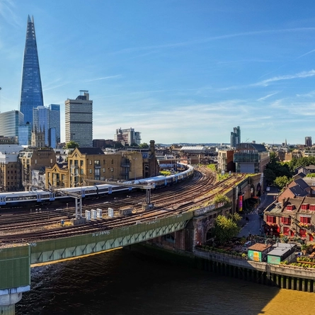 Panoramic view of London’s South Bank featuring The Anchor pub beside the River Thames, railway tracks and trains, with The Shard and the city skyline in the background.