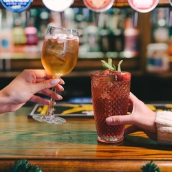 A hand picking up a cocktail served in a wine glass and topped with mini marshmallows, from a pub bar. On the bar is another cocktail served in a highball glass and garnished with mint, another hand holding the glass.