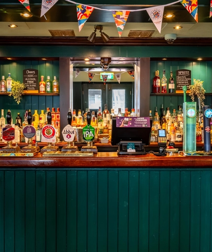 A close up view of the bar and beer taps at the Royal George in Euston; with golden drinks trays on the counter, and bottles of alcoholic spirits on illuminated wooden shelves behind the bar.