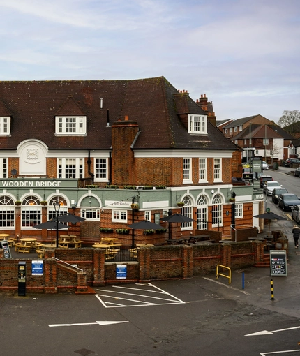 The exterior facade, signage, and beer garden seating area of the Wooden Bridge in Guildford, with wooden picnic tables and shade umbrellas.