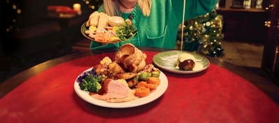 A roast dinner featuring roast meat, Yorkshire pudding, vegetables and gravy served on a plate, sits on a table with a plate of Christmas pudding and custard. A woman is sitting at the table holding a plate of prawn cocktail in one hand and a jug of custard in the other, and pouring it over the Christmas pudding.