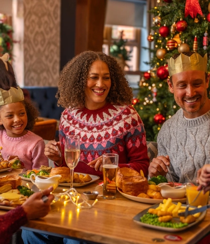 A family enjoys a festive Christmas meal at a dining table beside a decorated Christmas tree adorned with red and gold baubles, candy cane ornaments and warm fairy lights. Several diners wear paper crowns from Christmas crackers while sharing food and conversation. The table is set with traditional roast dinners, gravy boats, glasses of orange juice, sparkling drinks and Christmas crackers. Seasonal decorations and warm lighting create a cosy Christmas restaurant setting.