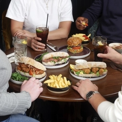 A group of four people seated around a small round wooden table, sharing a meal. The table is filled with plates of food, including baguette-style sandwiches, a burger, and several small bowls of chips. There are also drinks on the table, including glasses of soda with straws and a cup of coffee. Only the individuals’ torsos and arms are visible as they sit closely together, reaching toward the food.