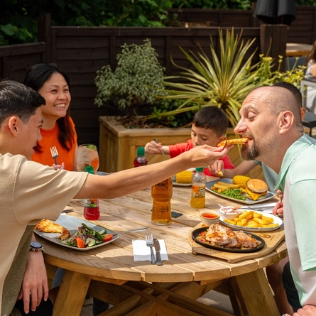 A family seated at an outdoor table in a beer garden, enjoying a meal, a youth is feeding his dad a bite of his pizza..