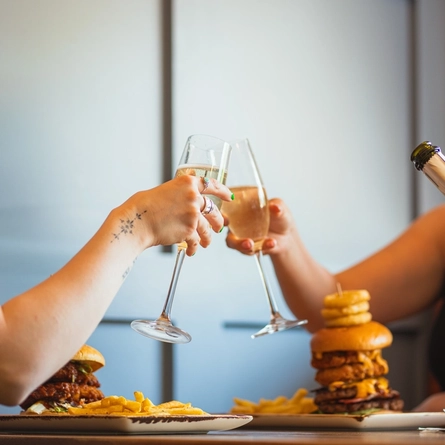 A lifestyle image of people sitting at an indoor table enjoying a burger and a glass of sparkling wine.