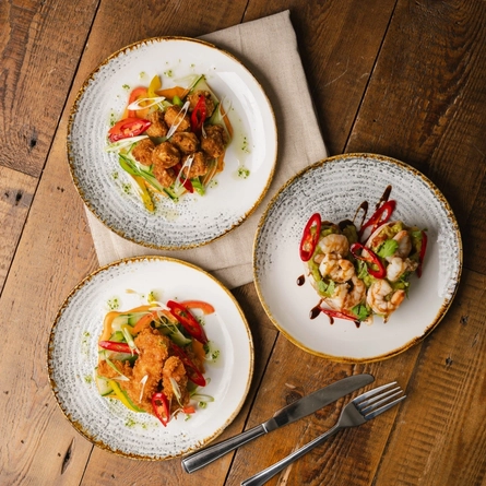 A bird's eye view of three plates of food sitting on a cloth napkin on a wooden restaurant table, along with a knife and fork.