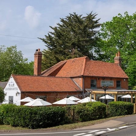 A view from across the road of the exterior facade, signage, and beer garden seating area of the Ye Olde Leathern Bottel in Barkham, with shade umbrellas and string lights. 