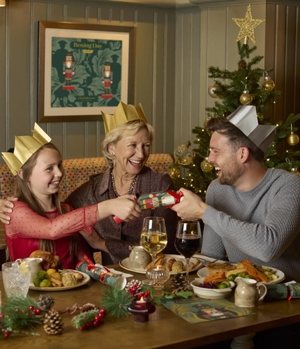 An image showing 2 adults and a child sat within the interior restaurant seating area pulling a cracker while enjoying Christmas Day dishes at a Chef & Brewer venue.