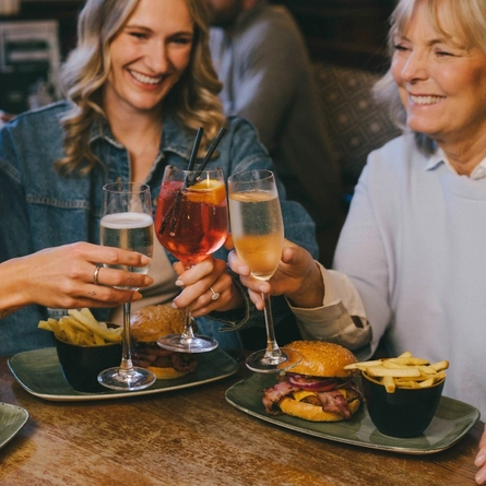 A lifestyle image focusing on 3 friends sat at a table enjoying bottomless burger mains and various drinks, celebrating with a cheers within the interior restaurant seating area at a Greene King venue.