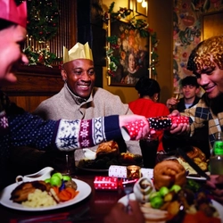 A group of people are gathered around a table enjoying a festive meal. They are wearing paper crowns, and the table is set with plates of food. Two individuals in the foreground are pulling a Christmas cracker. The background is decorated with Christmas decorations. 