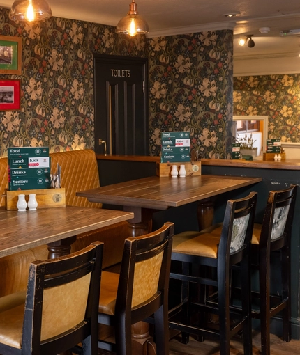 An interior restaurant seating area at The Royal Oak; with an upholstered booth seat along one side of the room, and framed artwork on the floral wallpapered wall above it.
