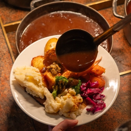 An over the shoulder view of a person standing at a carvery counter, holding a plate loaded with new potatoes, roast potatoes, mashed potato, vegetables, stuffing, and Yorkshire pudding, while using a ladle to scoop gravy out of the large roasting dish on the counter in front of them and drizzle it over the plate.
