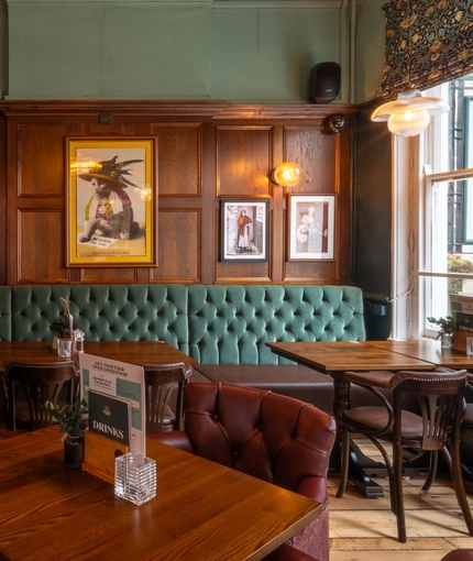 An interior restaurant seating area at The Plough, with upholstered chairs and booth seats, and framed artwork on the wood panelled wall.