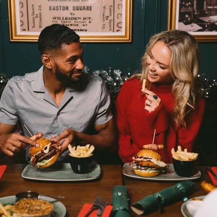 Two people sitting at a restaurant table with plates of burgers and chips in front of them. One person is picking up their burger, the other person is holding a chip, and a Christmas garland lit with fairy lights is visible in the background.