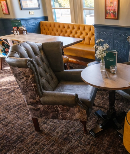 A close up view of a table in a corner of The Talbot Inn, with two upholstered wingback chairs, a pouffe, and a drinks menu on the table next to a vase of flowers and a tealight in a glass.