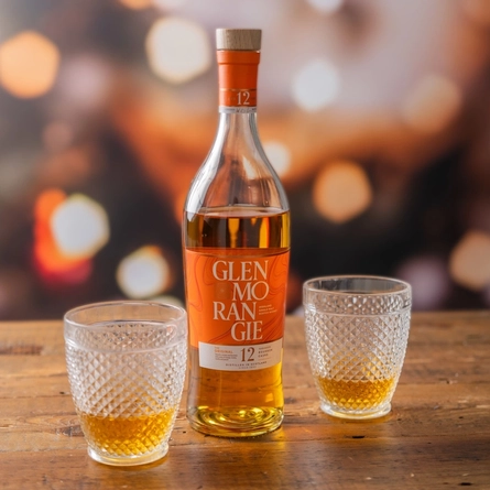 A bottle of February's Malt Of The Month - Glenmorangie whisky. The bottle stands on a wooden table, in between two glasses of the whisky.