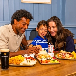 Two adults and a child are seated at a wooden table in a pub, enjoying a midweek meal. The table is filled with dishes including tacos, chips, corn on the cob and breaded bites, alongside a pint of Guinness, a glass of cola with lemon and a children's juice carton. Blue panelled walls and wooden seating create a relaxed pub setting in the background.