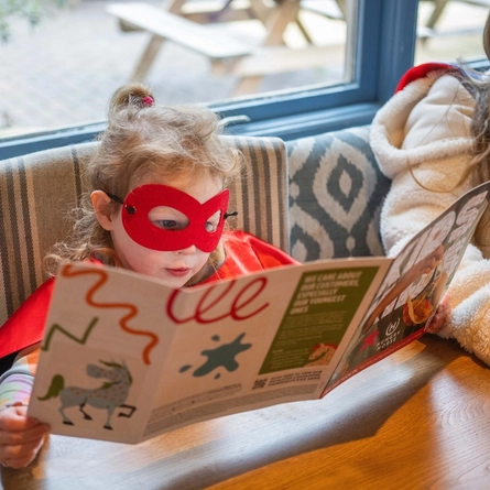 Two children dressed as superheros sat at an indoor wooden table, reading a cgildren's comic, activity book.