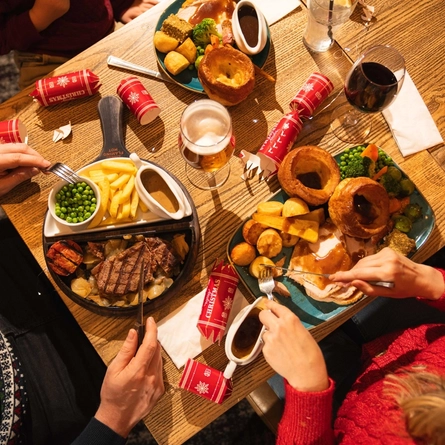Overhead view of a festive dining table set with Christmas meals and drinks in a pub. Plates include a traditional roast dinner with turkey, Yorkshire puddings, roast potatoes, vegetables and gravy, alongside a steak meal with chips, peas and grilled tomato. Glasses of lager and red wine, Christmas crackers and shared tableware complete the festive dining scene.
