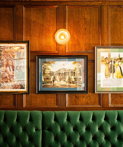 A close up view of framed artwork on a wood panelled wall inside The Plough. A green, upholstered booth seat is visible below the pictures.