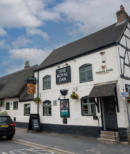 The exterior facade and signage of The Royal Oak in Bishops Cleeve; with a thatched roof, and flower baskets hanging on the walls.
