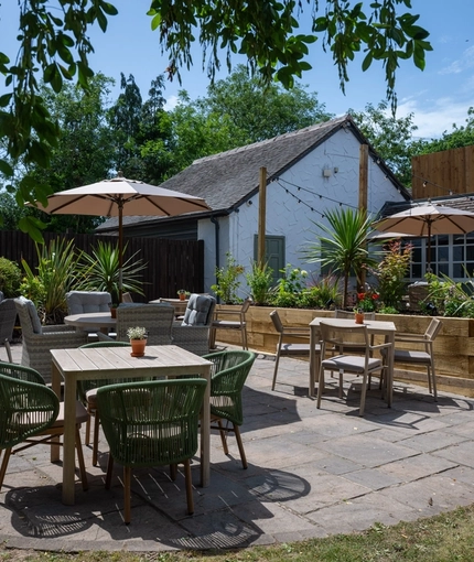 The paved exterior beer garden seating area at The Rising Sun in Wistaston, with shade umbrellas on some of the tables, a wooden planter box filled with plants and flowers, and string lights overhead.