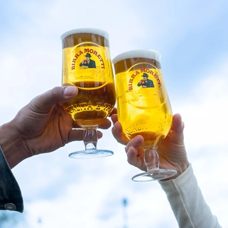 Two hands holding aloft a pint of Birra Moretti each against the sky as a backrop.