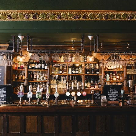 A close up view of the wood panelled bar inside the Masons Arms in Mayfair, with glasses hanging in racks above the counter, and grape vines carved into the coving above.