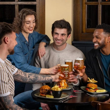 Four individuals sit around a table in a restaurant or pub setting. Three of them are seated, each holding a drink in a toast. The fourth individual stands beside the table, raising a wine glass. On the table are plates with burgers, bowls of fries, and small dipping containers.