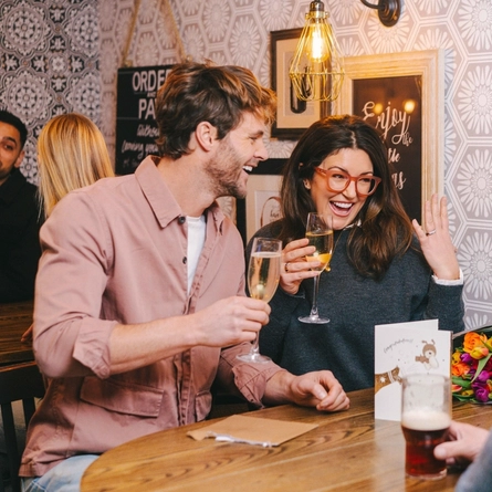 A newly engaged couple sitting in the restaurant and seating area with friends and drinks, in a Greene King pub showing the engagement ring.