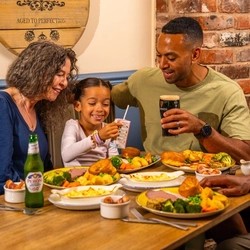 Three people seated around a wooden table in a pub, enjoying Sunday roast meals with Yorkshire puddings, roast potatoes and vegetables. A child holds a juice carton while an adult holds a pint of Guinness. An alcohol-free beer bottle and dishes of cauliflower cheese and pigs in blankets are on the table. The setting features blue panelled walls, exposed brickwork and a wooden barrel-style sign reading “The Mill House”.